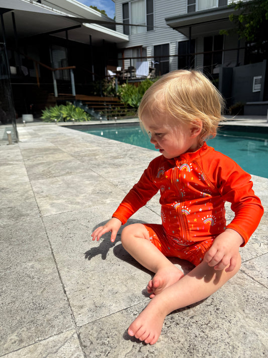 Child in an orange rainbow swimsuit sitting by a pool